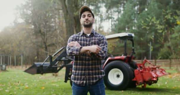 Joven granjero sonriendo mirando directamente a la cámara se para cerca del tractor — Vídeos de Stock