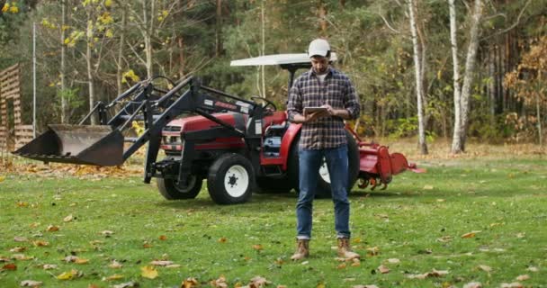 Een jonge man gebruikt een tablet, die op herfstdag bij een landbouwmachine staat — Stockvideo