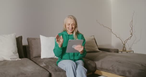 Femme âgée souriante commençant un appel vidéo sur une tablette assise sur un canapé — Video