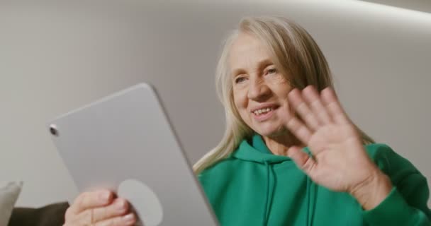 한연로 한 여자가 소파에 앉아 있는 태블릿으로 비디오 통화를 시작하면서 웃고 있다 — 비디오
