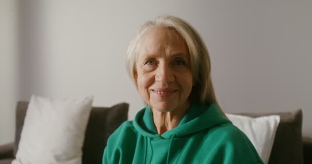 Mulher idosa sorrindo enquanto olha para a câmera, sentada no sofá, close-up — Vídeo de Stock