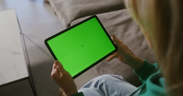 Donna anziana sorridente che inizia una videochiamata su un tablet seduta su un divano — Video Stock