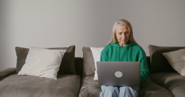 Uma mulher idosa sorri quando usa um laptop, sentada em um sofá — Vídeo de Stock