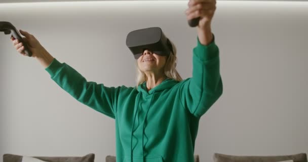 Μια ηλικιωμένη γυναίκα που φοράει γυαλιά εικονικής πραγματικότητας παίζει online χρησιμοποιώντας joysticks — Αρχείο Βίντεο