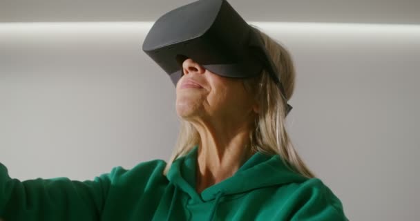 Een oudere vrouw met een virtual reality bril speelt online met joysticks — Stockvideo