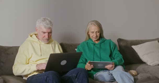 Un anciano usa una computadora portátil mientras está sentado junto a una mujer mayor usando una tableta — Vídeo de stock