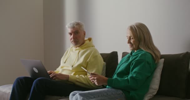 Un anciano usa una computadora portátil mientras está sentado junto a una mujer mayor usando una tableta — Vídeo de stock