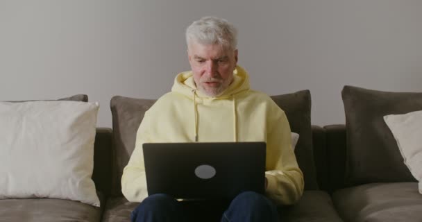 Ένας ηλικιωμένος άντρας χαμογελάει αρχίζει να κάνει βιντεοκλήση, χαιρετώντας τον καθισμένο σε έναν καναπέ στο σπίτι — Αρχείο Βίντεο