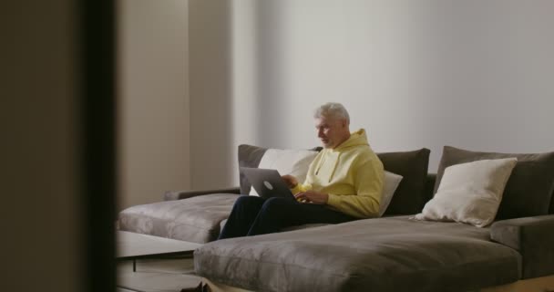 An elderly man smiling starts video call, waving hello sitting on a sofa at home — Stock Video