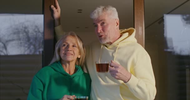 Um casal de idosos bebe chá e conversa, de pé no terraço em uma noite fria — Vídeo de Stock