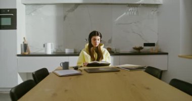 Kulaklıklı genç bir kadın kitap okuyor ve evde müzik dinliyor.