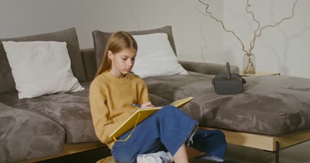 Девушка делает заметки в блокноте, сидя на полу рядом с диваном дома — стоковое видео