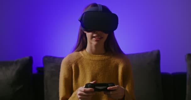 Ένα κορίτσι με γυαλιά εικονικής πραγματικότητας, παίζει ένα online παιχνίδι, κάθεται στον καναπέ — Αρχείο Βίντεο