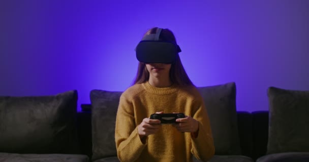 Seorang gadis dengan kacamata virtual reality, bermain game online, duduk di sofa — Stok Video
