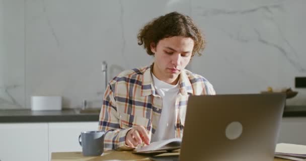 Een jongeman gebruikt een laptop, zittend aan een bureau, gelegd door vele boeken en notitieboekjes — Stockvideo