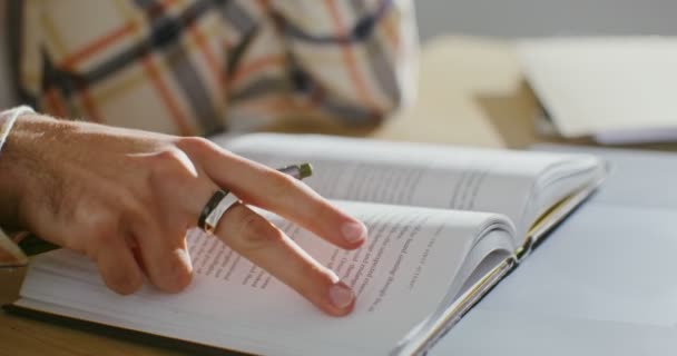 Un hombre gira una hoja de un libro sentado en una mesa con un bolígrafo en la mano de cerca — Vídeo de stock