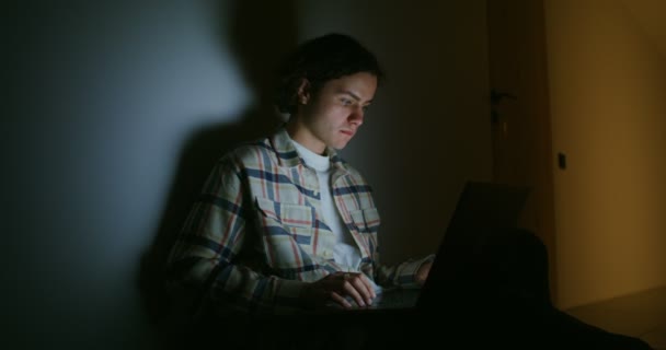Bir adam dizüstü bilgisayar kullanıyor. Sırtı alacakaranlıkta duvara dönük oturuyor. — Stok video
