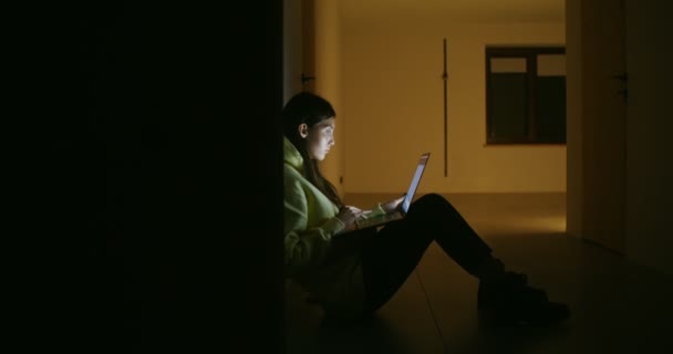 A woman sits in the darkness on the floor in back against wall and uses a laptop — Stock Video