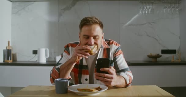 Молодой человек пьет кофе, не отрывая глаз от экрана мобильного телефона — стоковое видео