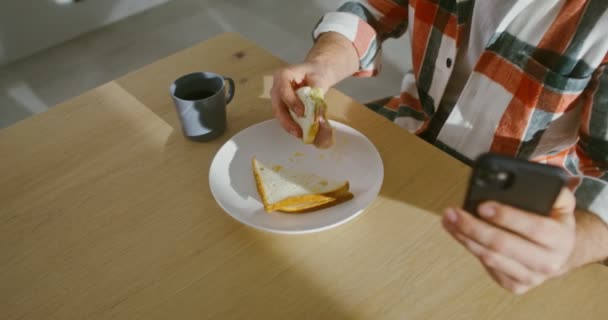 Een man is aan het ontbijten terwijl hij in de keuken zit en gebruikt een mobiele telefoon — Stockvideo