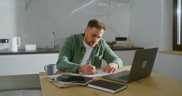 A man with a serious look works at a laptop, sitting in a modern kitchen — Vídeo de Stock