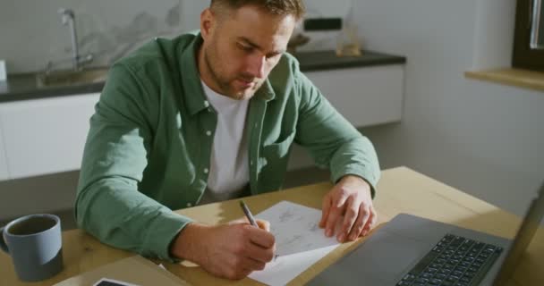 A man with a serious look works at a laptop, sitting in a modern kitchen — Stock Video