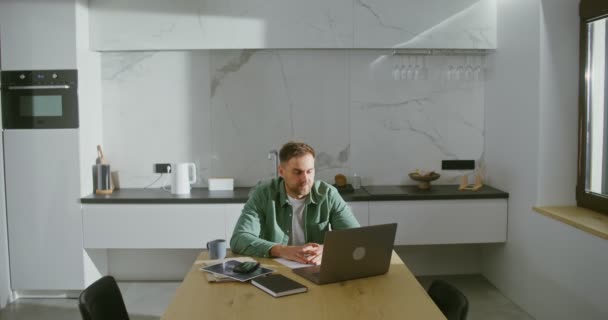 Nuori mies puhuu videolinkillä kannettavalla tietokoneella, istuu keittiön pöydässä — kuvapankkivideo
