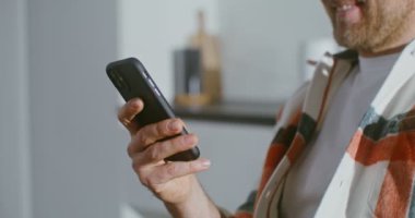Genç adam telefon ekranına bakarken gülümsüyor, mutfakta oturuyor.