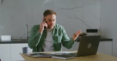 Adamın biri cep telefonuyla konuşuyor. Mutfak masasında dizüstü bilgisayarın önünde oturuyor.