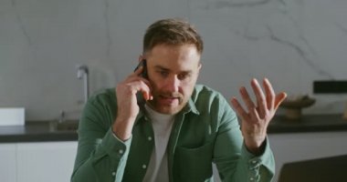 Adamın biri cep telefonuyla konuşuyor. Mutfak masasında dizüstü bilgisayarın önünde oturuyor.