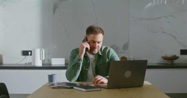 ノートパソコンの前のキッチンテーブルに座ってる男が携帯電話で話してる — ストック動画