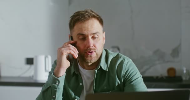 Mężczyzna rozmawia przez telefon komórkowy, siedzi przy kuchennym stole przed laptopem — Wideo stockowe