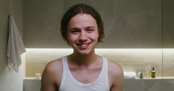 Νεαρός άνδρας χαμογελά κοιτάζοντας κατευθείαν στην κάμερα ενώ στέκεται στο μπάνιο — Αρχείο Βίντεο