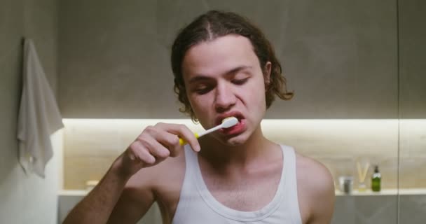 Young man squeezes toothpaste onto a toothbrush and begins to brush his teeth — Stockvideo