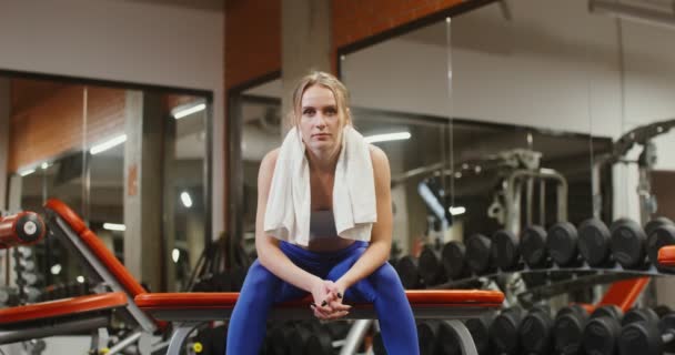 Mujer joven mira directamente a la cámara sentado en el banco de gimnasia en el gimnasio — Vídeos de Stock