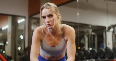 Genç kadın doğrudan kameraya bakar spor salonundaki jimnastik masasında oturur.