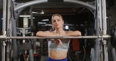 Genç bir kadın kameraya bakar spor salonunda egzersiz makinesine yaslanır.