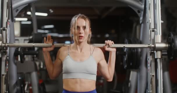 Jonge vrouw quats met een lange halter met gewichten met behulp van een trainingsmachine — Stockvideo