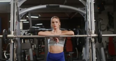 Genç bir kadın kameraya bakar spor salonunda egzersiz makinesine yaslanır.