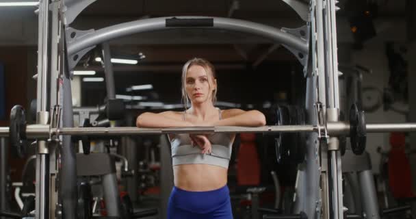 Uma jovem mulher olha para a câmera, apoiando-se em uma máquina de exercícios o ginásio — Vídeo de Stock
