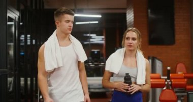 Genç bir adam ve kadın modern bir spor salonunda koşu bandına yürürken konuşuyorlar.