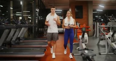 Genç bir adam ve kadın modern bir spor salonunda koşu bandına yürürken konuşuyorlar.