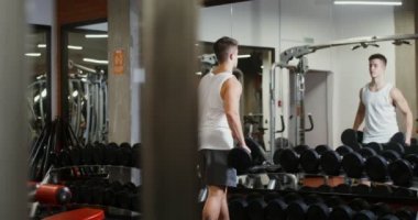 Bir adam eline dambıl alır ve spor salonundaki aynaya bakarak antrenman yapmaya başlar.