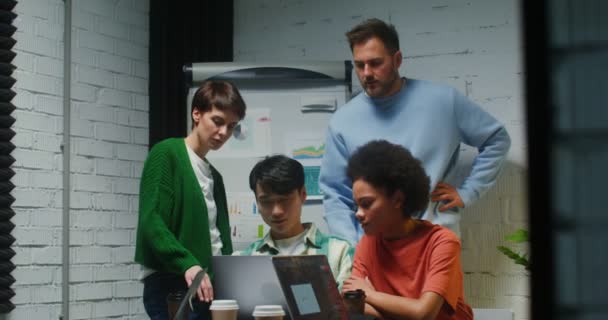 Группа людей стоит рядом с рабочим местом своего коллеги и смотрит на его ноутбук — стоковое видео