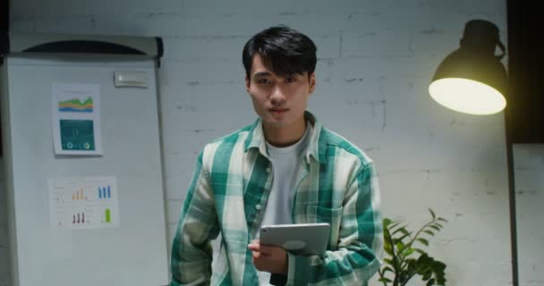 Young Asian man smiling looking at camera while standing with tablet in office — Stockvideo