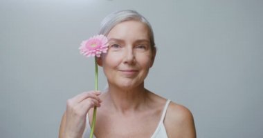 Yaşlı bir beyaz model Gerbera çiçeğiyle poz veriyor, yüzünü okşuyor.
