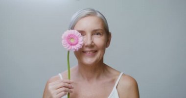 Yaşlı bir beyaz model Gerbera çiçeğiyle poz veriyor, yüzünü okşuyor.