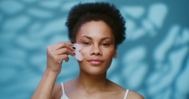 En ung afroamerikansk kvinna som masserar ansiktet med Gua Sha — Stockvideo