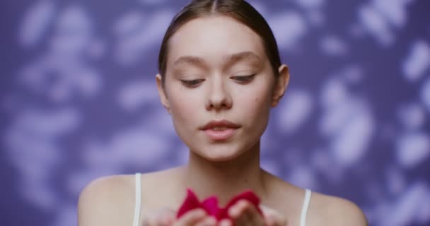 Una joven modelo sopla los pétalos de rosa acostada en sus palmas y mira a la cámara — Vídeos de Stock