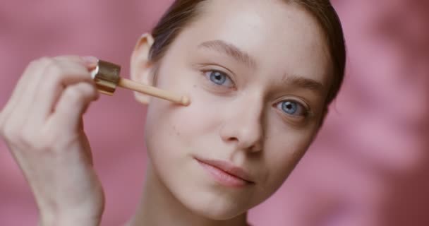 A young woman applying foundation with a dropper to her cheeks, close-up — ストック動画